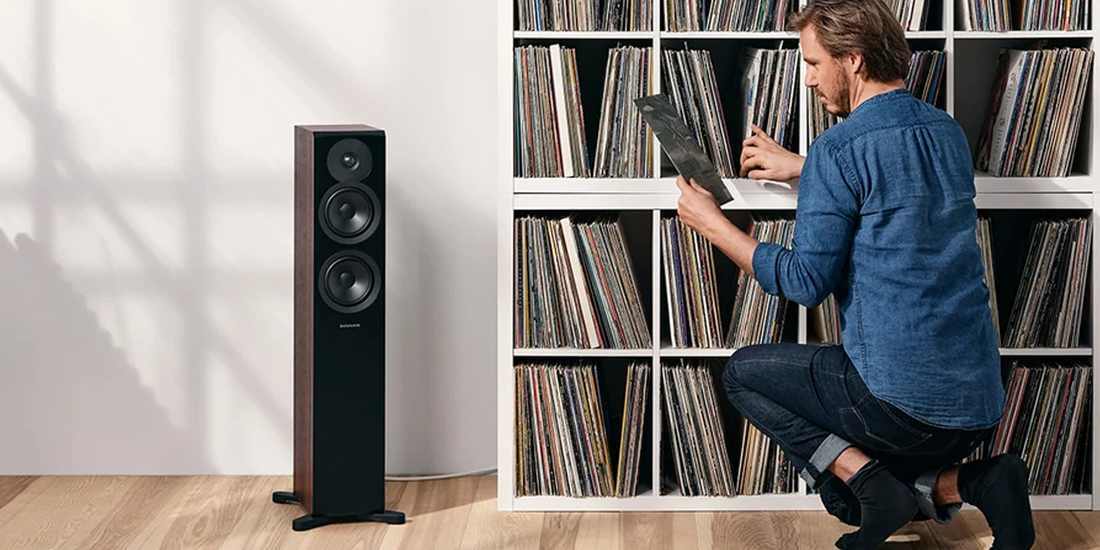 Pic showing a man kneeling at some shelves holding vinyl albums a Dynaudio Emit 30 speaker to his left